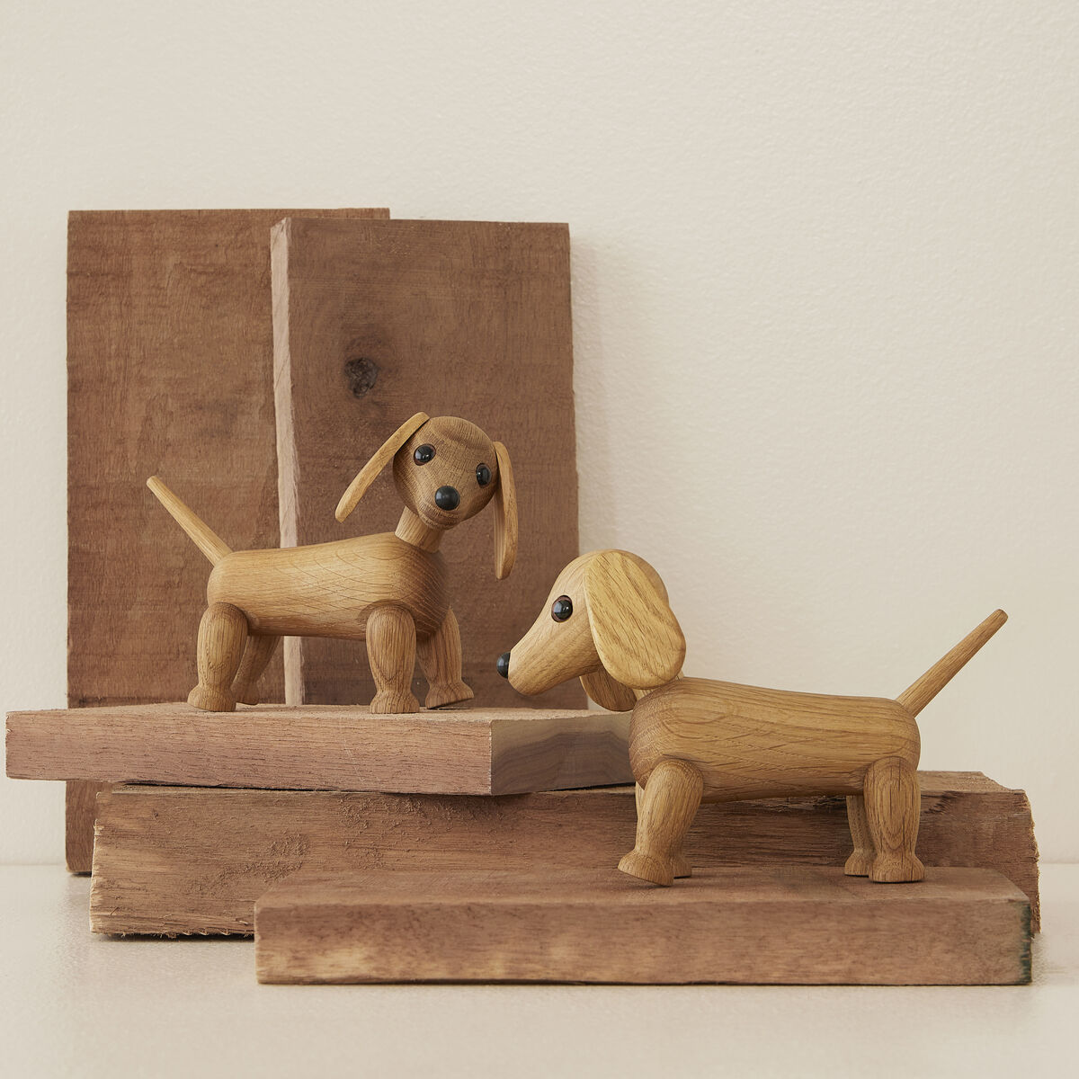 Zwei Holzfiguren in Dackelform aus hellem, naturbelassenem Holz stehen auf gestapelten Holzbrettern vor beiger Wand. Die Figuren sind detailreich mit langen Ohren, schwarzen Augen und charakteristisch ausgestreckten Schwänzen gestaltet.