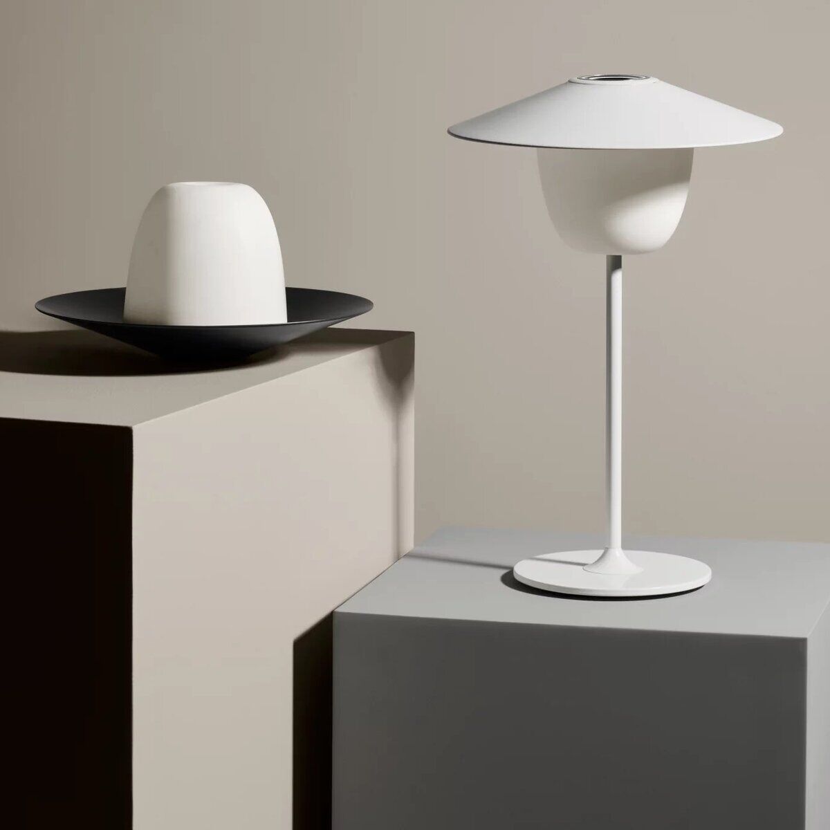 Eine mobile LED Tischleuchte namens ANI, die in einem minimalistischen, modernen Design gehalten ist. Die Lampe hat einen weißen Lampenschirm und einen schlanken, eleganten Fuß. Ein weiteres Modell hängt von der Decke, während ein kleinerer, zylinderförmiger Lampenschirm auf einem niedrigeren Podest steht. Die Farben im Bild sind hauptsächlich Weiß und Grautöne.