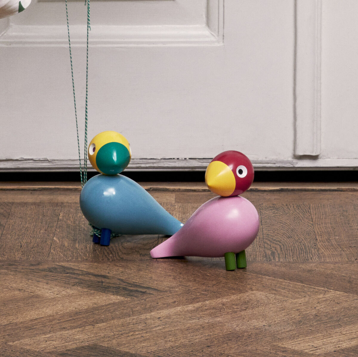 Ein Kinderzimmer mit einer Holzpferd-Wippe, einem geflochtenen Korb mit einer weißen Decke, die mit bunten Vogelmotiven bedruckt ist, bunten Luftballons und zwei farbenfrohen Holzvögeln 'Sunshine' von Kay Bojesen auf dem Holzboden. Im Hintergrund eine weiße Tür mit einer Dekotasche, ebenfalls mit bunten Vogelmotiven.