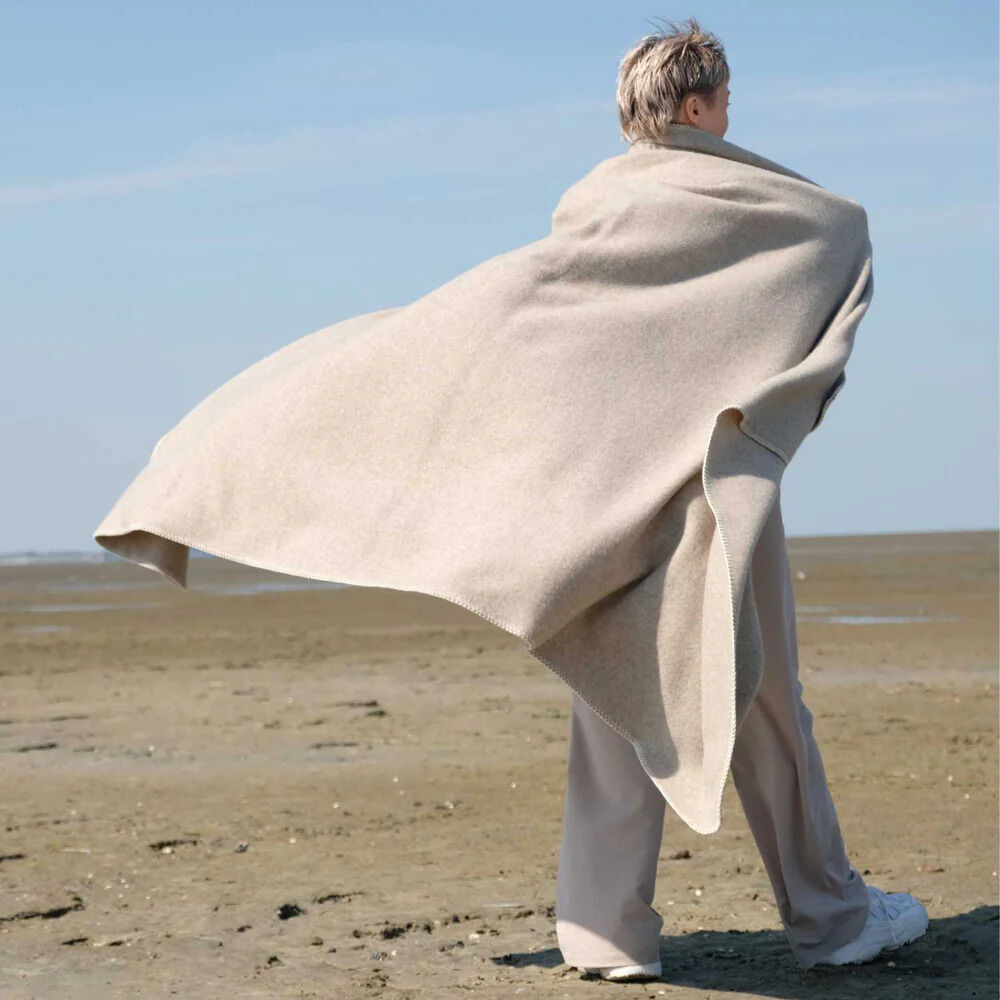 Eine Person mit heller, stilvoller Merinowolldecke „SkinGreyRock“, die locker um die Schultern gelegt ist, steht auf leicht sandigem, offenem Strand bei windigem Wetter. Die Decke wirkt groß, weich und hochwertig verarbeitet. Im Hintergrund erstreckt sich der flache Strand, Atmosphäre von Ruhe und Natur.