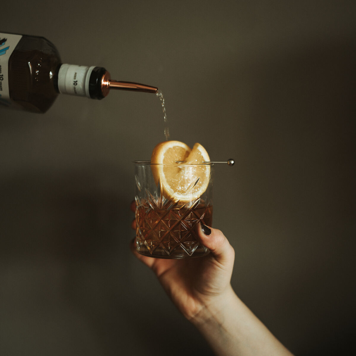 Eine Hand hält ein Glas mit einer Zitronenscheibe und Eiswürfeln, in das ein Rum namens »Ron Piet Rum«, 10 Jahre gereift, gegossen wird. Das Bild hat einen dunklen Hintergrund und vermittelt eine stilvolle und exklusive Atmosphäre.