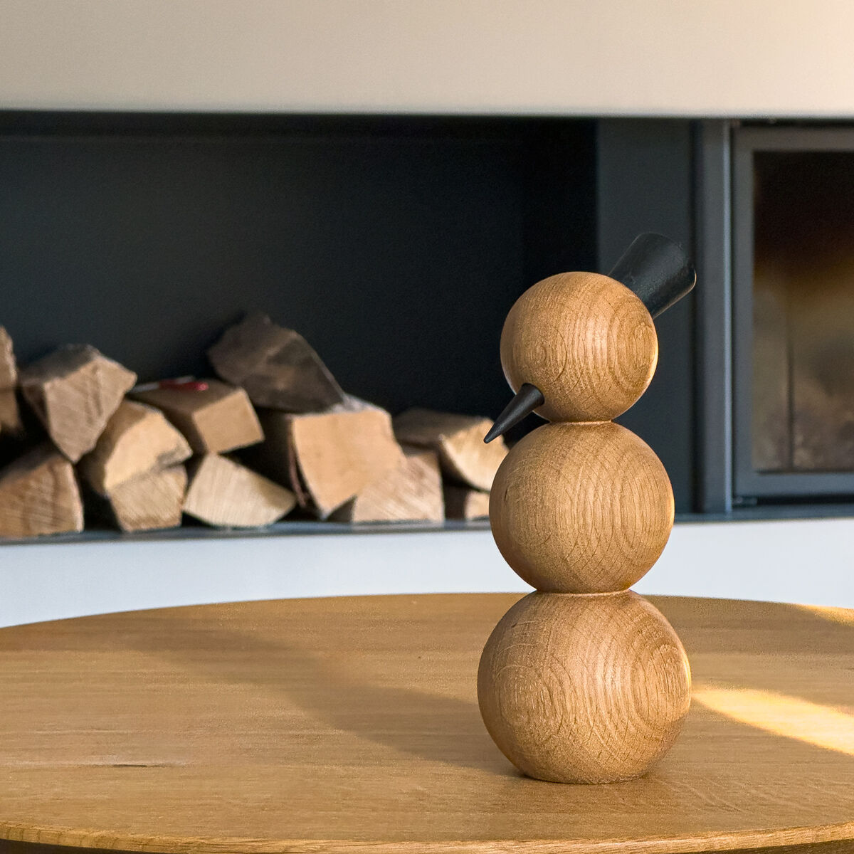 Reduzierte Holzfigur Schneemann aus warm brauner, deutlich gemaserter Esche auf einer hellen, spiegelnden Holzplatte. Zwei gedrechselte Kugeln bilden Kopf und Körper; schwarze, schräge Stahlrohre ragen seitlich am Kopf heraus. Die Platte spiegelt die Figur. Im Hintergrund liegen Scheite in einem dunklen Kaminfach.