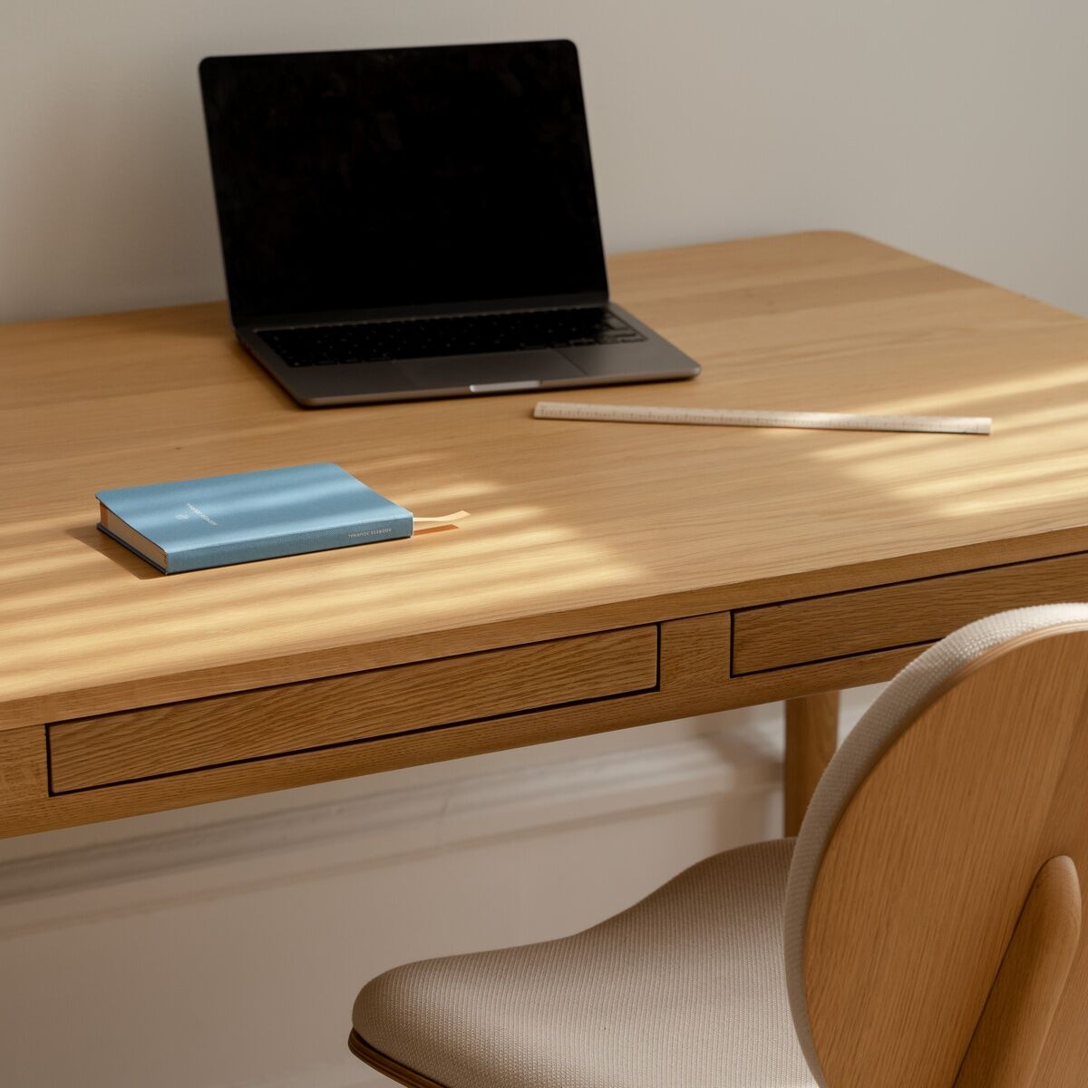 Heller, minimalistischer Schreibtisch »Heart'n'Soul« der Marke Umage aus Eiche mit zwei Schubladen. Auf dem Tisch stehen ein schwarzer Laptop, ein hellblaues Notizbuch und ein Stift. Passend dazu eine skandinavische, gepolsterte Holzstuhl in Beige.
