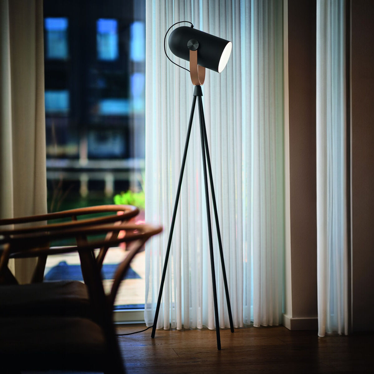 Eine moderne Stehleuchte der ZEIT Sonderedition »Carronade II« mit schwarzem Metallschirm und braunen Holzdetails in einem stilvollen Wohnzimmer mit Vorhängen im Hintergrund.