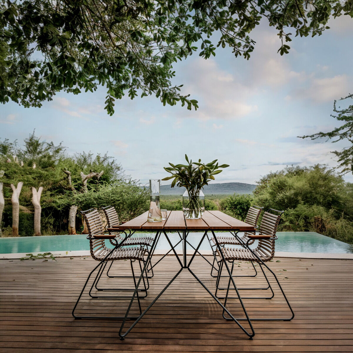 Ein moderner Gartentisch namens 'SKETCH' steht auf einer hölzernen Terrasse vor einem Pool mit üppiger Vegetation im Hintergrund. Der Tisch hat eine natürliche, braune Oberfläche mit einem schlanken, schwarzen Stahlrahmen. Einige passende Stühle aus Metall und Holz sind um den Tisch herum angeordnet. Die Szene vermittelt einen Eindruck von Ruhe und Naturgenuss.