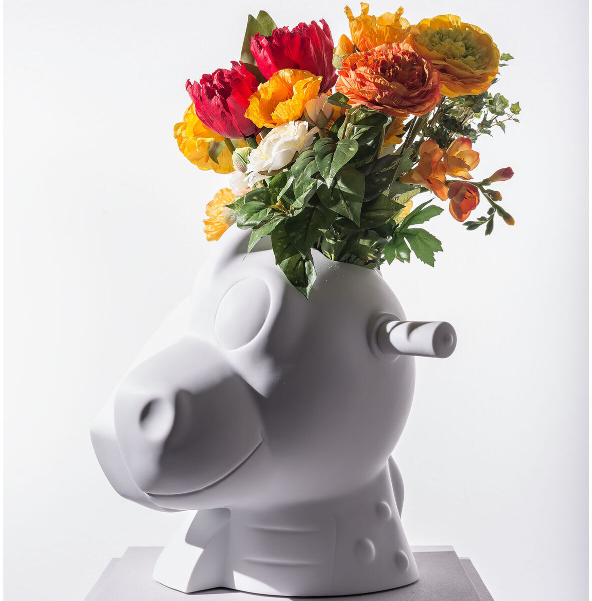 Die Skulptur »Split-Rocker« von Jeff Koons aus dem Jahr 2012 zeigt ein weißes, stilisiertes Pferde- oder Dinosaurierkopfobjekt, das als Blumentopf verwendet wird. Bunte Blumen in Gelb, Orange, Rot und Grün ragen aus der Oberseite der Skulptur heraus. Die Skulptur ist bekannt für ihren modernen und künstlerischen Touch und steht auf einer weißen Plattform.