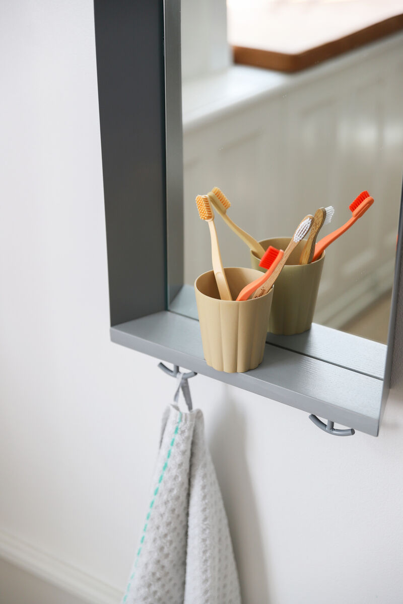 Ein Wandspiegel für Kinderzimmer, mit integriertem Regal. Auf dem Regal stehen vier Zahnbürsten in einem beige Zahnbürstenhalter. Darunter hängt ein Handtuch. Der Spiegel und das Regal sind modern und funktional gestaltet in Grautönen.