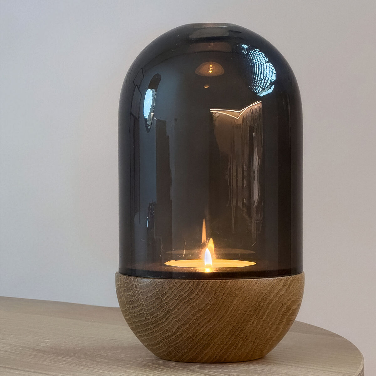 Windlicht »GLOW« der Marke finbeaux auf einem Holztisch. Der obere Teil besteht aus Rauchglas in Glockenform, darunter ein runder Holzsockel. Im Inneren brennt eine Kerze und erzeugt ein warmes, stimmungsvolles Licht. Die Kulisse ist minimalistisch und modern gestaltet.