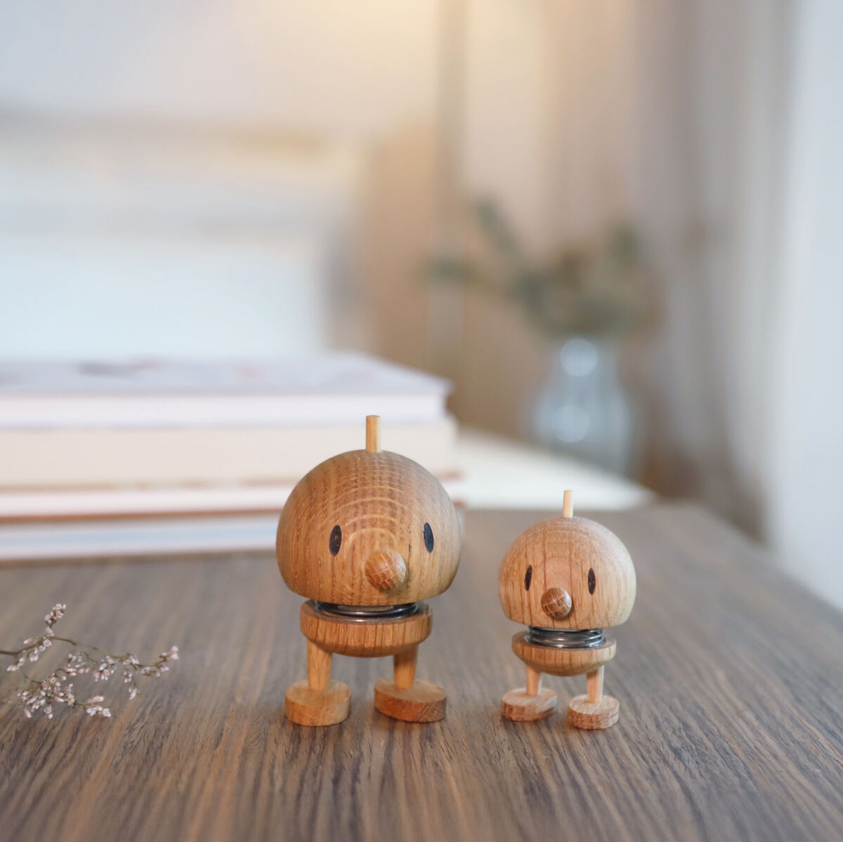 Zwei hölzerne Hoptimisten in verschiedenen Größen aus Eiche stehen auf einem Tisch vor einem hellen, unscharfen Hintergrund mit einem Bücherstapel. Die Figuren wirken freundlich und dekorativ, ideal als Geschenk oder Dekoration für moderne Wohnräume.