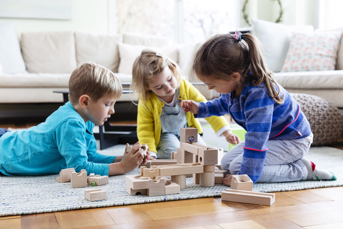 Drei Kinder spielen auf einer Bodenmatte mit einem Kugelbahn Starter-Set aus Holz. Das Set besteht aus mehreren Bauteilen, die zu einer Bahn zusammengesetzt werden können. Es sind helle Holztöne und bunte Kinderkleidung zu sehen.