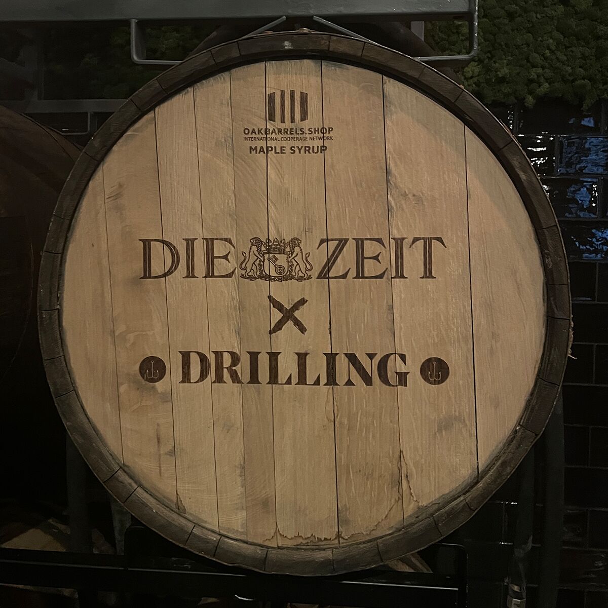 Großes, rundes Eichenfass mit der Aufschrift 'DIE ZEIT x DRILLING', umgeben von dunklen Fliesen und rustikalem Ambiente. Das Fass dient als Ankündigung für eine exklusive Whiskey-Sonderedition und steht für hochwertige Handwerkskunst und Genusskultur.