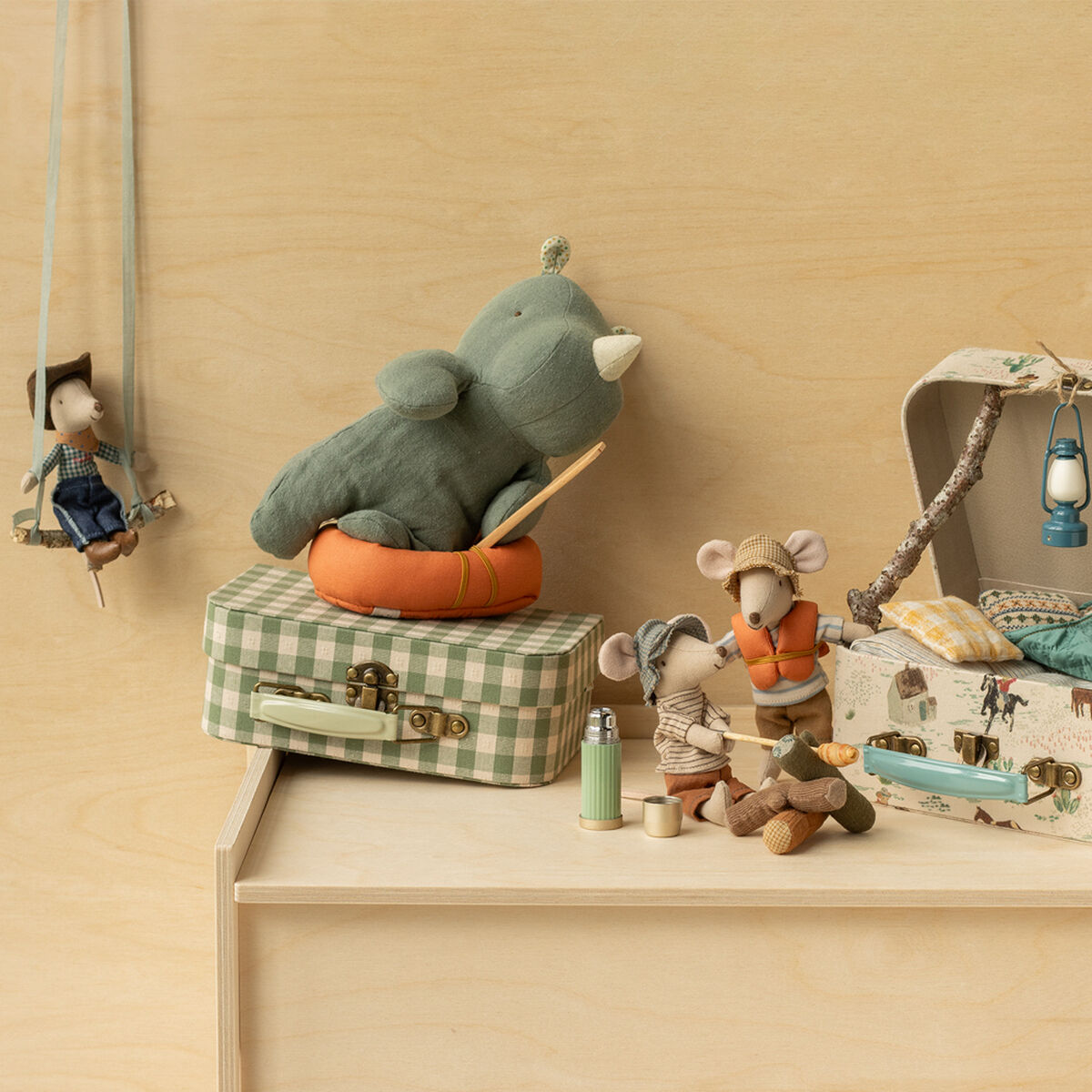 Mehrere liebevoll gestaltete Stoffmäuse und ein grünes Stoffnashorn sitzen und liegen auf einem Holztisch und kleinen Koffern in naturnahen Farben. Eine Maus trägt eine Wanderkluft und ein Fernglas, rundherum befinden sich Camping-Deko wie Schlafsack, Laterne und Lagerfeuer. Die Szene ist gemütlich und lädt zum fantasievollen Spielen ein.