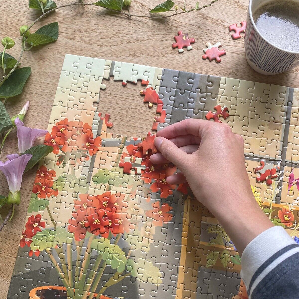 Eine Hand setzt ein Teil zu einem fast fertigen 500-Teile-Puzzle »Blossom« von VISSEVASSE zusammen. Das Puzzle zeigt rote Blumen in einem Topf auf einem Tisch. Daneben liegen Blüten und eine Tasse Kaffee auf hellem Holztisch.