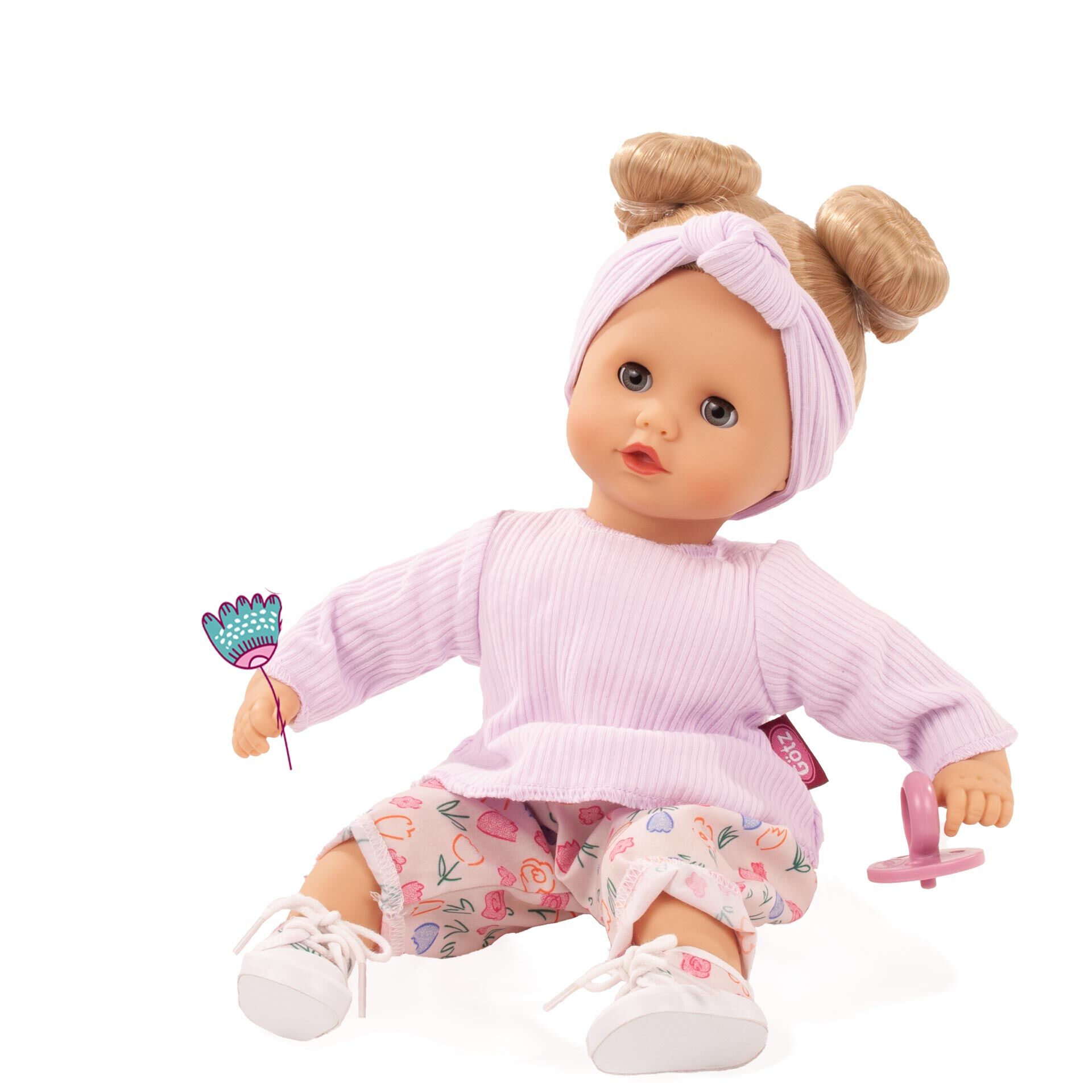 Eine niedliche Puppe namens »Muffin BliBlaBlume« sitzt auf dem Boden. Sie trägt ein fliederfarbenes Shirt, eine passende Schleifenstirnband und gemusterte Hosen in Rosa mit Blumenmotiven. Die Puppe hält ein kleines Blümchen und einen Schnuller in den Händen. Ihre Füße sind mit weißen Schuhen bekleidet. Geeignet als Spielzeug für Kinder.