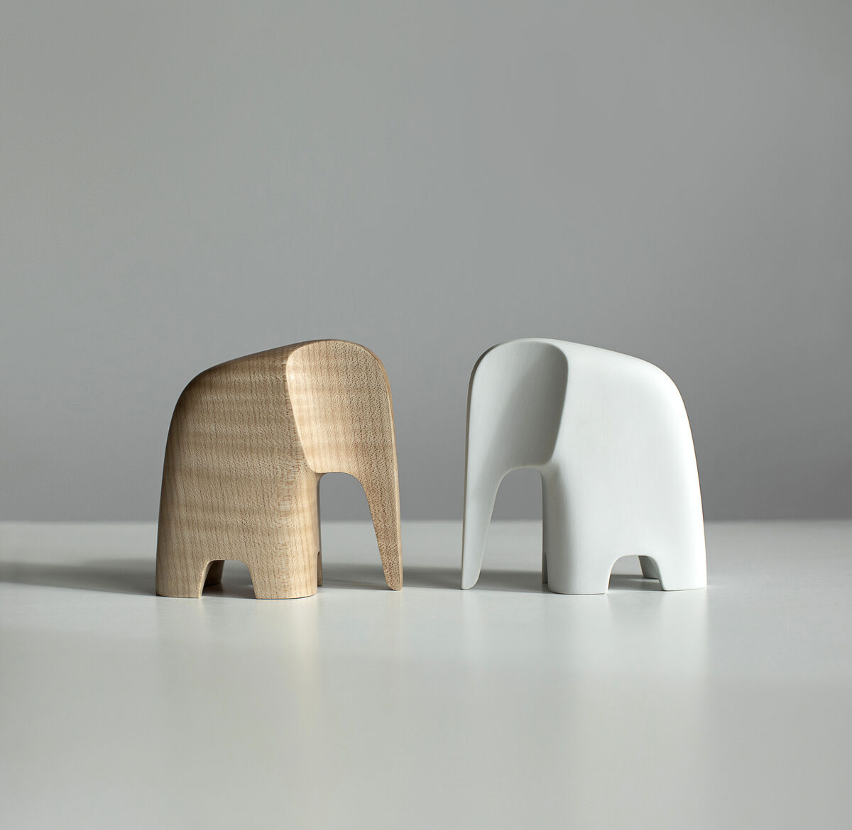 Zwei minimalistische Elefantenfiguren aus Holz und Keramik stehen nebeneinander auf einer weißen Oberfläche. Eine Figur ist naturfarbenes Holz, die andere ist weiß bemalt. Die Skulpturen haben ein modernes und schlichtes Design.