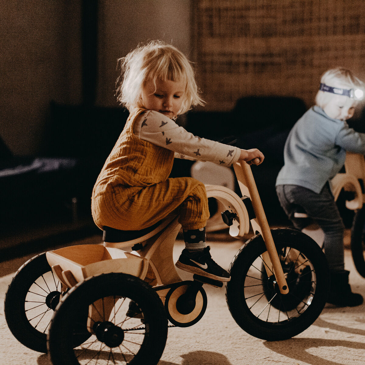 Zwei kleine Kinder spielen drinnen auf holzfarbenen Dreirädern, die als Erweiterung für Balance Bikes dienen. Das linke Kind trägt einen gelben Overall und das rechte nutzt eine Stirnlampe. Die Szene findet in einem gemütlichen, gedämpft beleuchteten Raum statt.