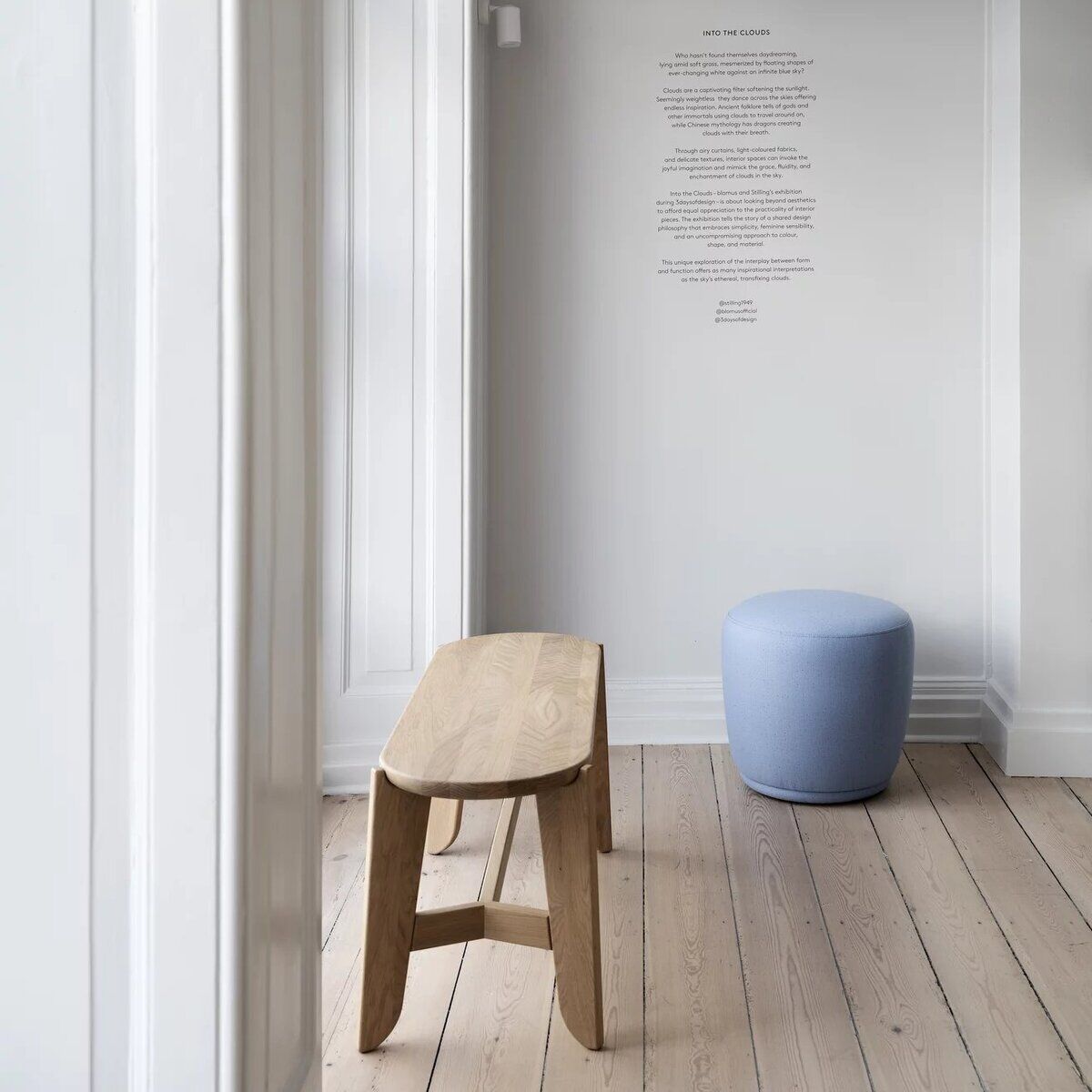 Eine minimalistische Sitzbank aus hellem Holz steht in einem modernen, hellen Raum mit Holzboden. Im Hintergrund befindet sich ein hellblauer Hocker und eine weiße Wand mit Text. Das Design vermittelt ein skandinavisches, schlichtes Ambiente.