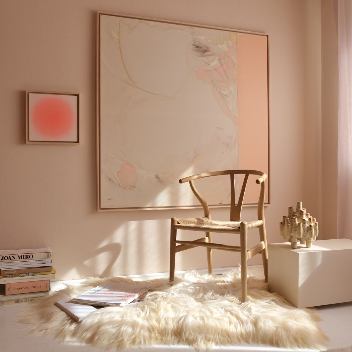 Quadratisches Kunstwerk in Apricot- und Pastelltönen hängt an einer hellen Wand, daneben ein minimalistischer Stuhl aus Holz, ein flauschiger Teppich und Bücher, ideal für moderne Wohnräume.