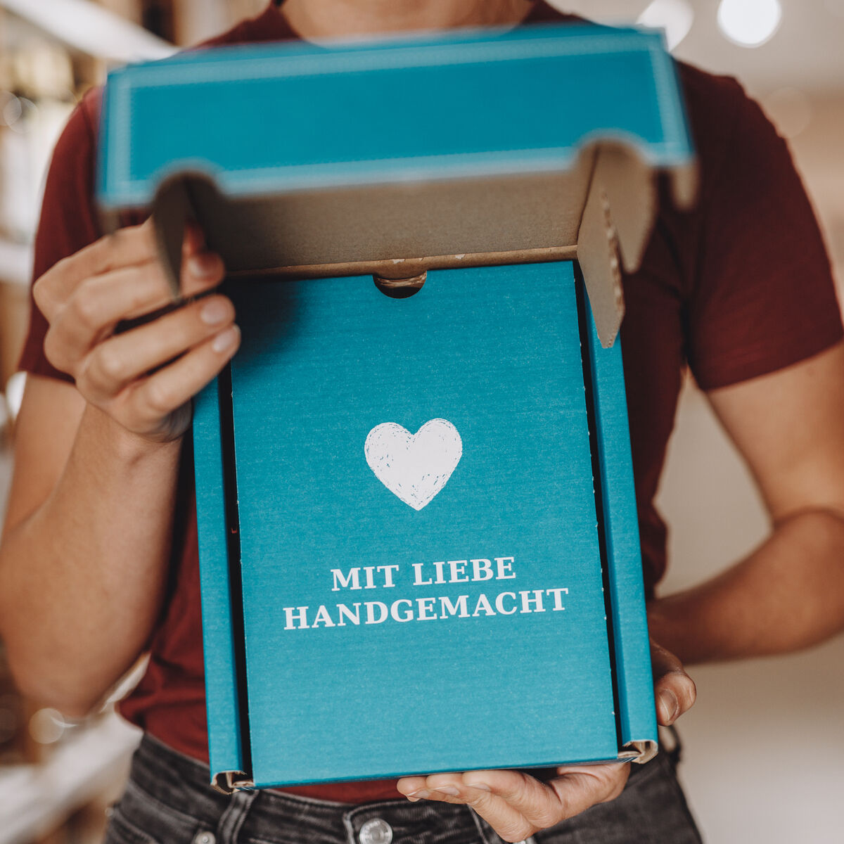 Eine Person hält einen geöffneten, türkisfarbenen Geschenkkarton mit weißem Herzsymbol und der Aufschrift 'Mit Liebe handgemacht' in den Händen. Der Karton ist Teil eines Geschenksets, das als Grillöl-Set mit dem Titel 'Grillmeisters Lieblinge' verwendet wird. Der Hintergrund ist unscharf und vermittelt eine freundliche Atmosphäre in einem modernen Umfeld.