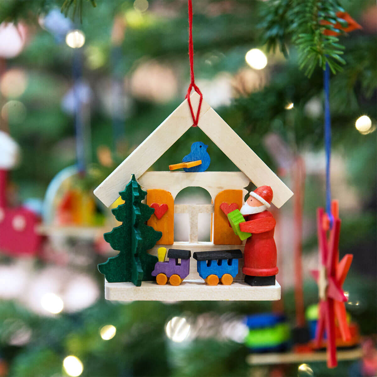 Ein dreiteiliges Geschenk-Set aus Holz-Weihnachtsschmuck-Anhängern in Form von Weihnachtsmannhäusern. Jedes Haus ist detailliert bemalt, hängt an einem Weihnachtsbaum und zeigt einen Weihnachtsmann, einen kleinen Weihnachtsbaum und bunte Fenster. Der Hintergrundausschnitt zeigt einen verschwommenen Weihnachtsbaum, der mit weiteren Schmuckstücken und Lichtern dekoriert ist.