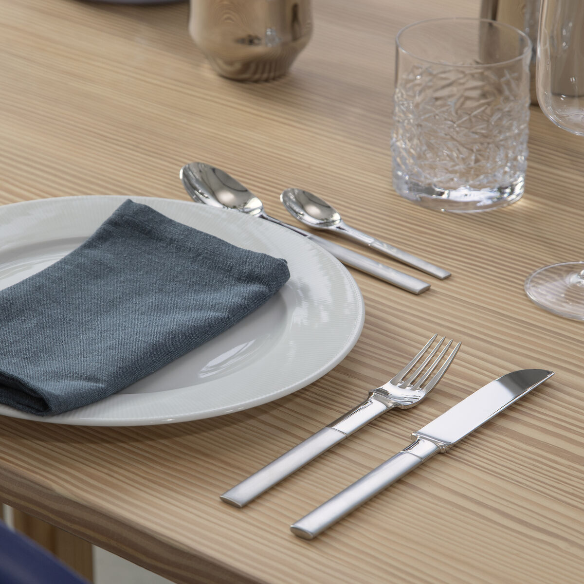 Ein gedeckter Tisch mit einem eleganten Besteckset. Silbernes Besteck liegt auf einem Holztisch neben einem weißen Porzellanteller und einem grauen Stoffserviette. Im Hintergrund befinden sich ein Wasserglas, ein Weinglas und eine silberne Vase mit Blumen.