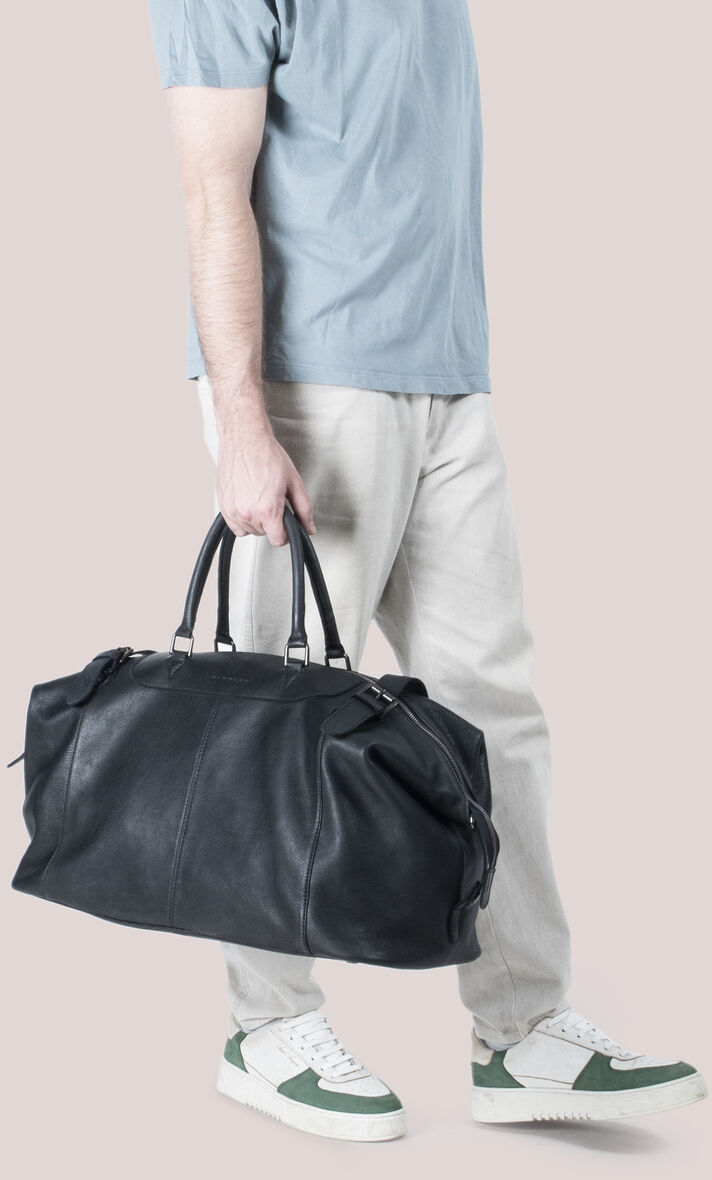 Eine hochwertige schwarze Reisetasche aus Rindleder, die von einem Mann in einem grauen T-Shirt, weißen Hosen und weißen Turnschuhen mit grünen Akzenten getragen wird.