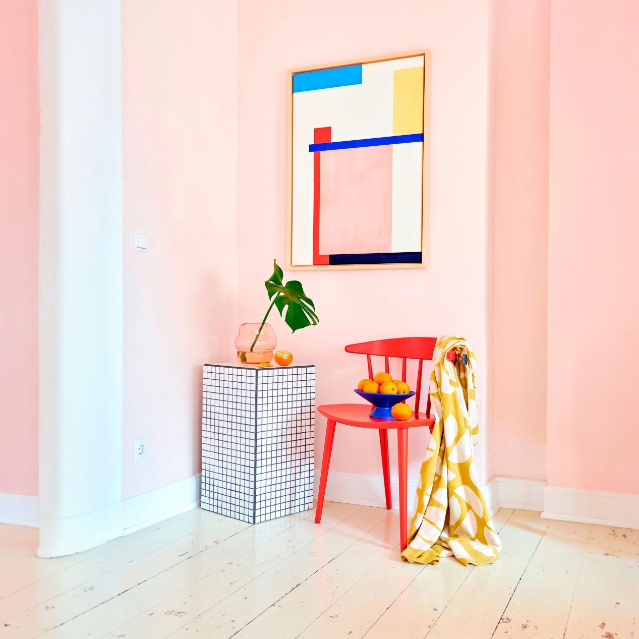 Leinwandbild »Fokke No 02« mit abstraktem, geometrischem Design in Blau, Rot, Gelb und Weiß an einer pastellrosa Wand, eingerahmt von moderner Wohnzimmereinrichtung mit rotem Stuhl, gemustertem Beistelltisch, Schale mit Orangen und dekorativem Blatt, helle und freundliche Atmosphäre.