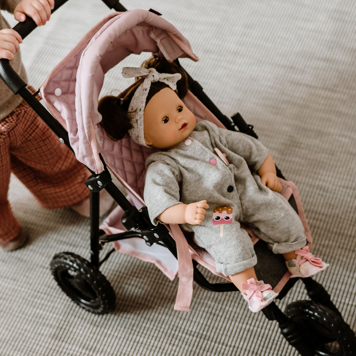 Ein Kleinkind schiebt eine Puppe namens »Maxy Muffin Popsicle« in einem rosa Puppenwagen auf einem hellen Teppich. Die Puppe trägt einen grauen Strampler und eine gemusterte Mütze. Das Bild vermittelt eine kindgerechte, spielerische Atmosphäre.