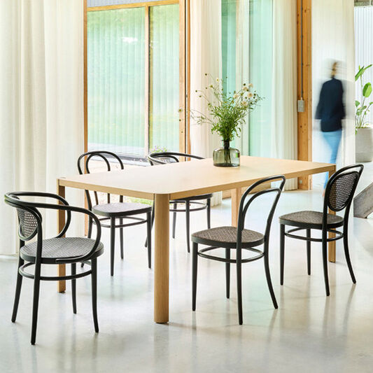 Im hellen Raum stehen vier schwarze Thonet Bugholzstühle »214« aus Buchenholz mit Rohrgeflecht um einen rechteckigen Holztisch. Auf dem Tisch steht eine Vase mit grünen Zweigen. Große Fenster und helle Vorhänge sorgen für viel Tageslicht, eine Person geht im Hintergrund.