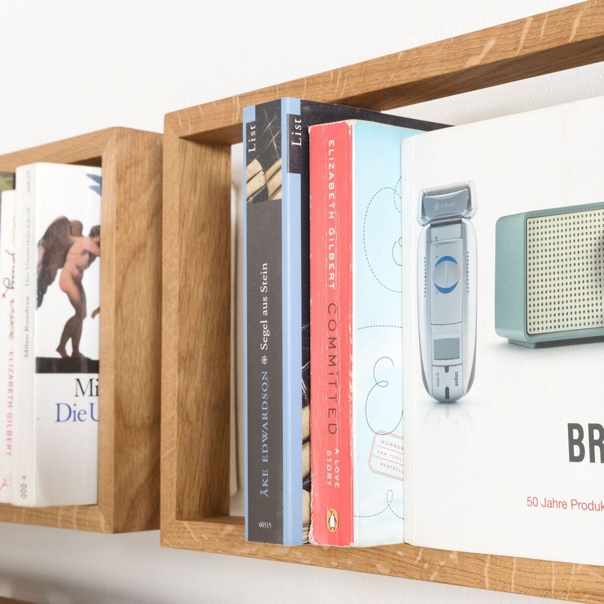 Ein Holzregal mit einer Reihe von sorgfältig arrangierten Büchern und anderen Gegenständen. Das Regal hat ein minimalistisches, modernes Design und besteht aus braunem Holz. Man sieht den Seitenteil und die obere Fläche des Regals, das an einer weißen Wand befestigt ist.