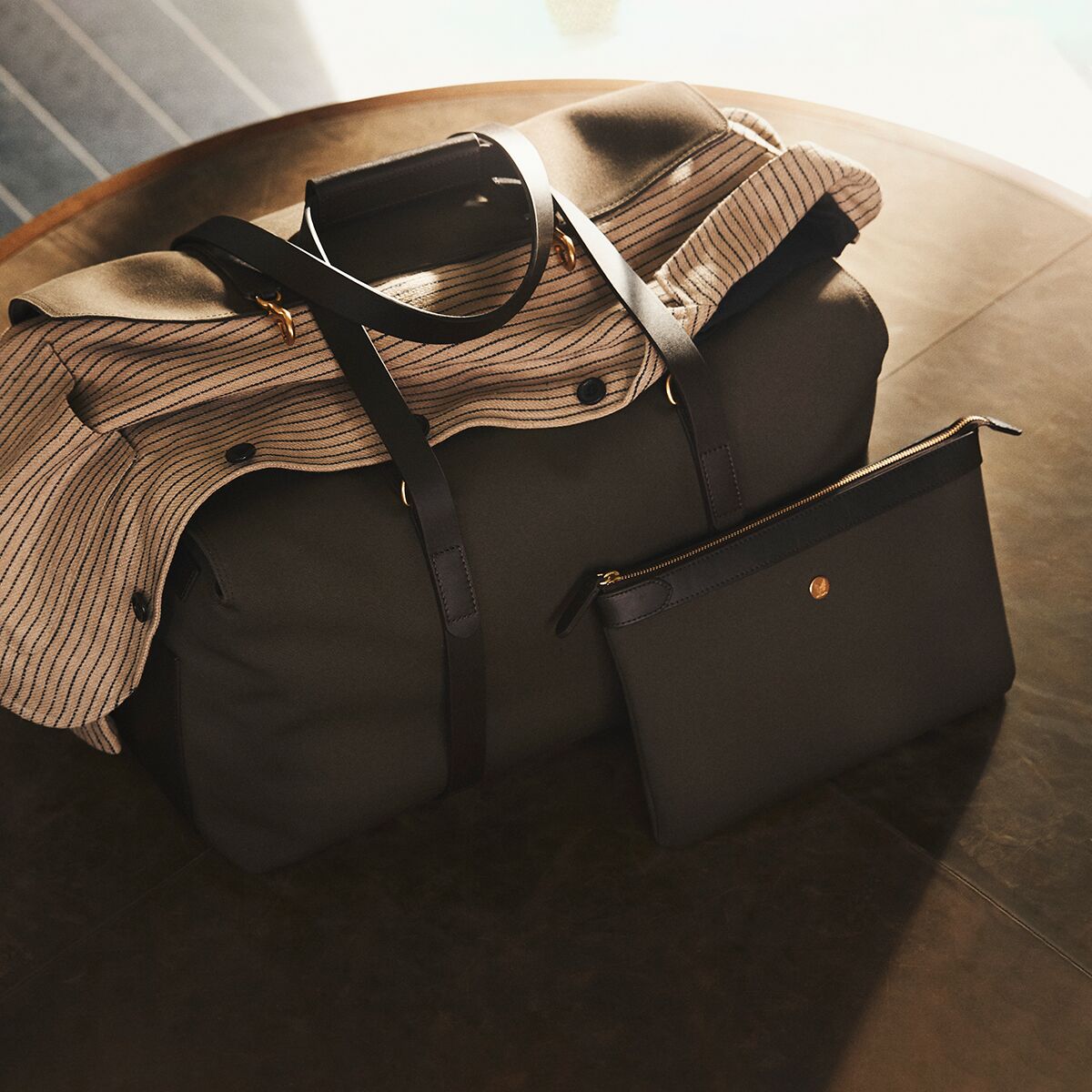 Auf einem edlen, dunklen Holztisch liegt eine große olivfarbene Tasche mit braunen Lederhenkeln und goldenen Details. Darauf platziert ist eine kleinere, passende Pouch »POUCH LARGE« von MISMO. Innen sind gestreifte Stoffelemente sichtbar, das Licht betont die hochwertigen Materialien und die elegante Verarbeitung.