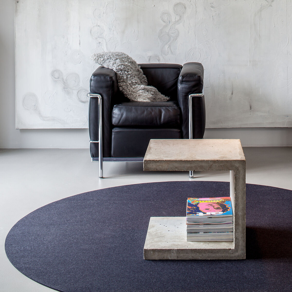 Ein eleganter Wohnbereich mit einem runden, schwarzen Teppich »Big Dot«. Im Hintergrund steht ein schwarzer Ledersessel neben einem modernen Beistelltisch mit Zeitschriften. Die Raumgestaltung ist minimalistisch und modern, geprägt von neutralen Farben und klaren Linien.