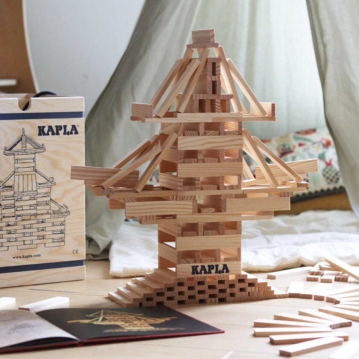 Ein kreatives Holzbaustein-Set bestehend aus vielen gleichmäßigen Holzplättchen, die zu einer komplexen Struktur zusammengebaut sind. Im Hintergrund stehen ein Karton mit Illustration und ein Buch mit Bauideen. Die gesamte Szene befindet sich in einem Kinderzimmer mit einer weichen Decke und Kissen im Hintergrund.