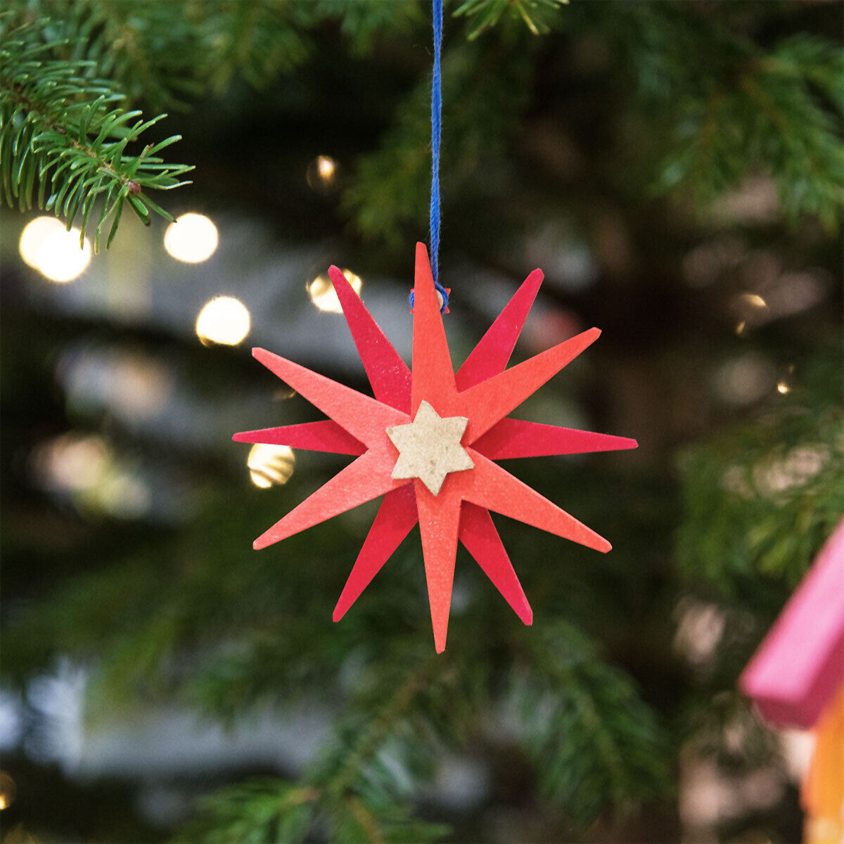 Ein rotes Weihnachtsornament in Sternform hängt an einem grün geschmückten Tannenbaum, beleuchtet von einer Lichterkette.