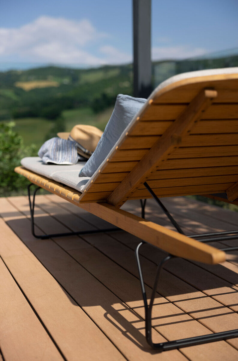 Liege »MOLO« aus Holz auf einer Terrasse, mit grauen Kissen und einem Strohhut, im Hintergrund eine grüne Landschaft und blauer Himmel.