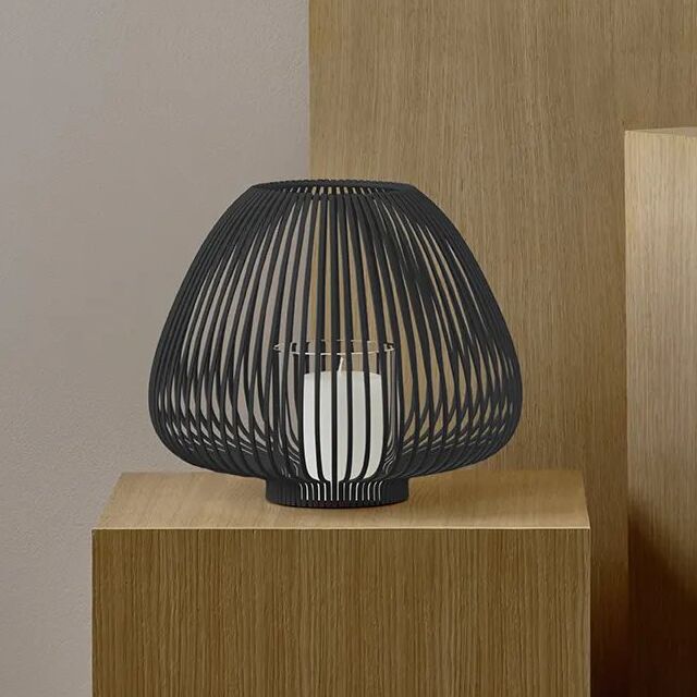 Moderne schwarze Laterne namens »LYVA« aus Metall mit rundem Design, steht auf einem hellen Holzpodest. Im Inneren der Laterne befindet sich ein weißes Kerzenglas. Die Szene ist minimalistisch und stilvoll arrangiert in einem hellen Raum.