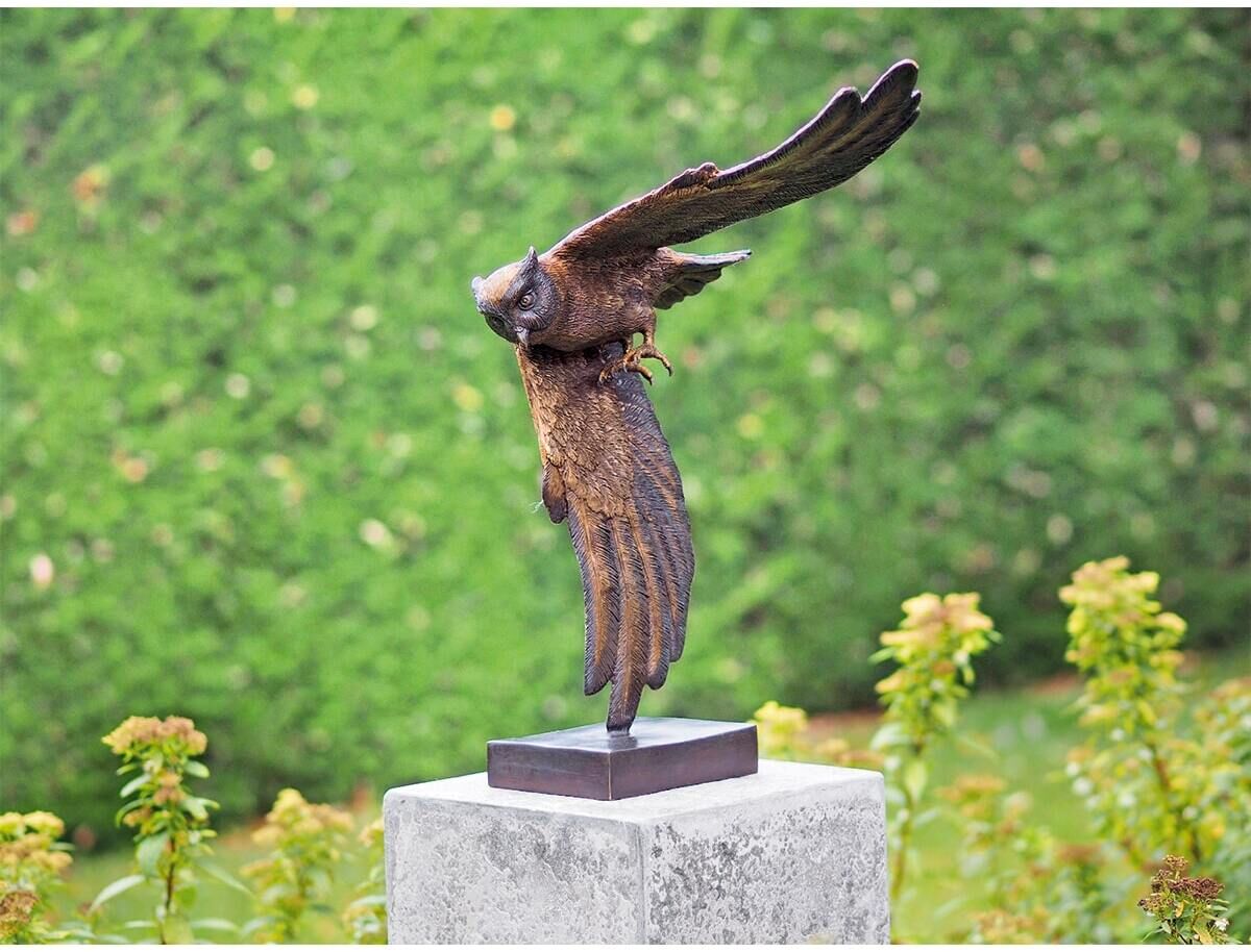 Metallskulptur einer fliegenden Eule, detailreich gestaltet, auf einem Sockel montiert, vor einem Hintergrund aus grünen Büschen.