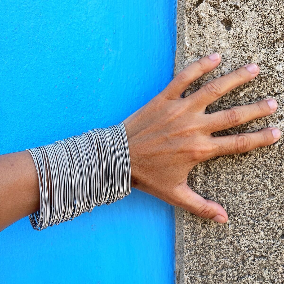 Eine Hand trägt ein Armband aus feinen Edelstahlfedern. Der Hintergrund ist zweigeteilt mit einem blauen und einem beigen Bereich.