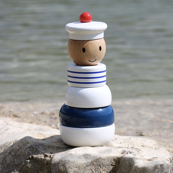 Maritime Stapelfigur aus Holz, die einen lächelnden Seemann mit blau-weißer Kleidung und rotem Hut darstellt. Die Figur sitzt auf einem felsigen Strand mit Meer im Hintergrund.