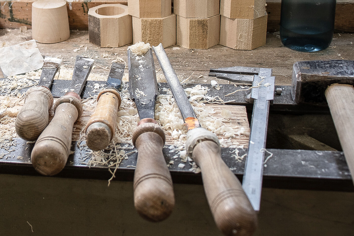 Holzmeißel mit abgenutzten Holzgriffen und ein Messschieber liegen auf einer Werkbank mit Holzspänen und Holzstücken.