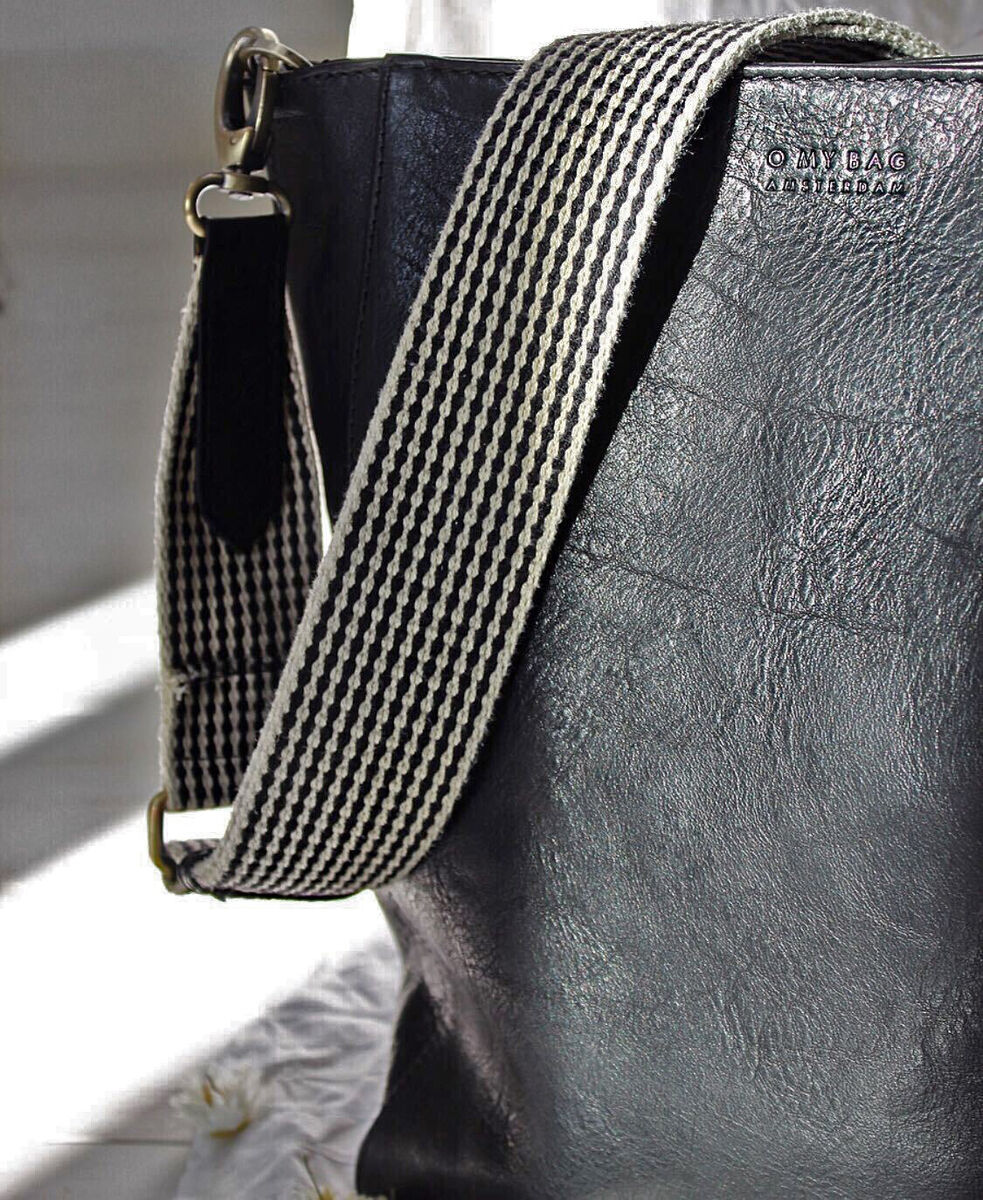 Schwarze Ledertasche mit breitem, weiß-schwarz gestreiftem Schulterriemen auf einer hellen Unterlage.