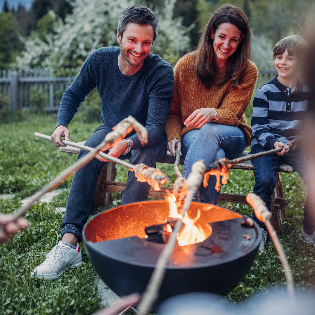 Eine Familie sitzt im Garten um eine Feuerschale vom Modell »BOWL 57« und röstet Marshmallows über dem offenen Feuer. Im Hintergrund sind Bäume und ein Gartenzaun zu sehen. Die Personen lächeln und genießen die gemeinsame Zeit im Freien.