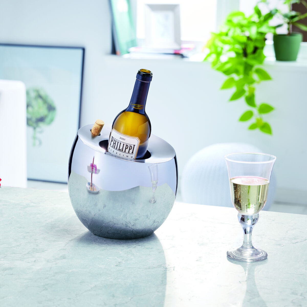 Ein silberner Weinkühler, der eine Weinflasche kühlt, steht auf einem modernen Küchentisch. Neben dem Weinkühler befindet sich ein Glas Weißwein und ein Serviertablett mit kleinen Kuchen. Im Hintergrund sind Pflanzen und Möbel zu sehen.