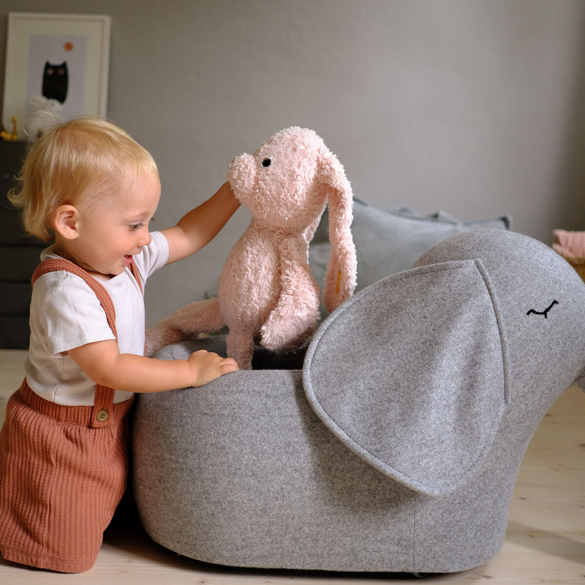Ein Kleinkind spielt mit einem weichen, grauen Elefanten-Spielzeug, das auf Rädern steht. Neben dem Elefanten befindet sich ein rosa Kuscheltier. Im Hintergrund ist ein Kinderzimmer zu sehen mit einem kleinen Regal.
