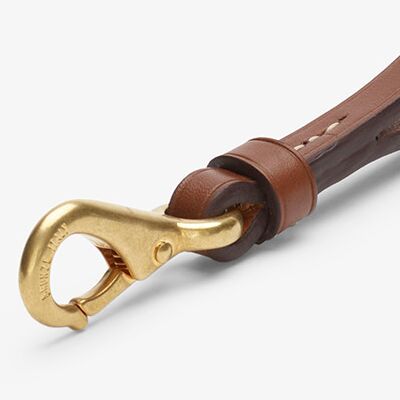 Detailaufnahme eines hochwertigen Schlüsselanhängers aus naturbraunem, pflanzlich gegerbtem Rindleder mit polierten Kanten und feinen Ziernähten. Ein massiver goldfarbener Karabinerhaken dient als Befestigung, Leder und Metallelement sind präzise verarbeitet und edel kombiniert.