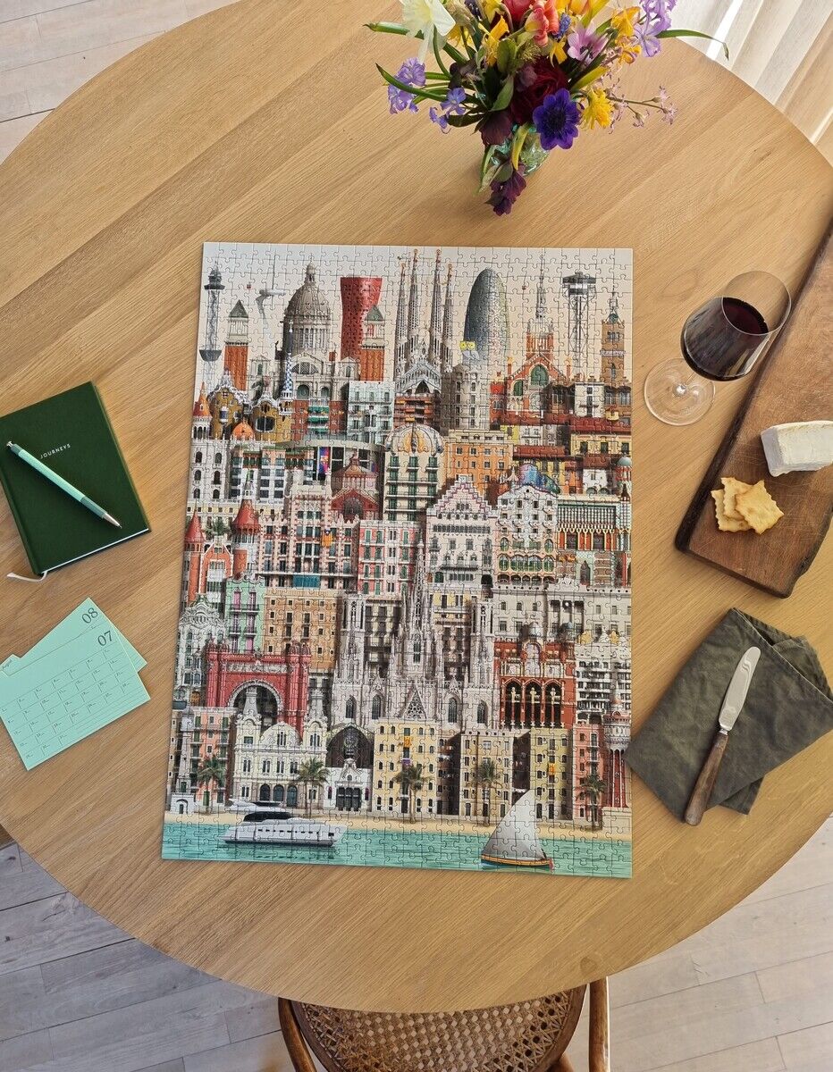 Ein 1000-Teile-Puzzle mit dem Motiv der Stadtansicht von Barcelona liegt auf einem Holztisch. Neben dem fertiggestellten Puzzle befinden sich eine Blumenvase, ein Glas Rotwein, ein Teller mit Käse, ein Notizbuch mit einem Stift und ein Kalenderblatt.