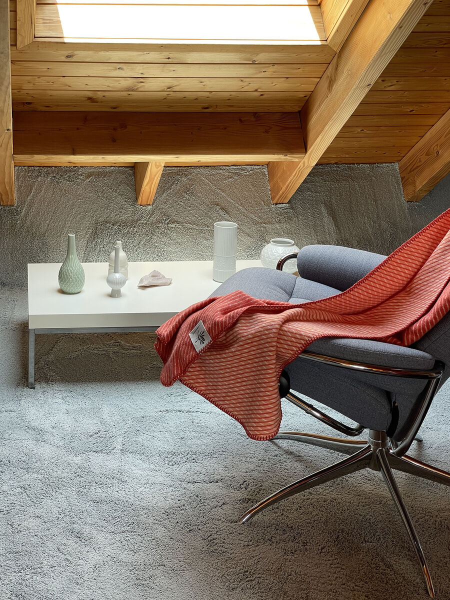 Eine rote Baumwolldecke mit diagonalem Muster ist über einen grauen Sessel drapiert. Der Sessel steht neben einem weißen Tisch mit Dekorationsgegenständen in einem hellen und modernen Dachgeschossraum.