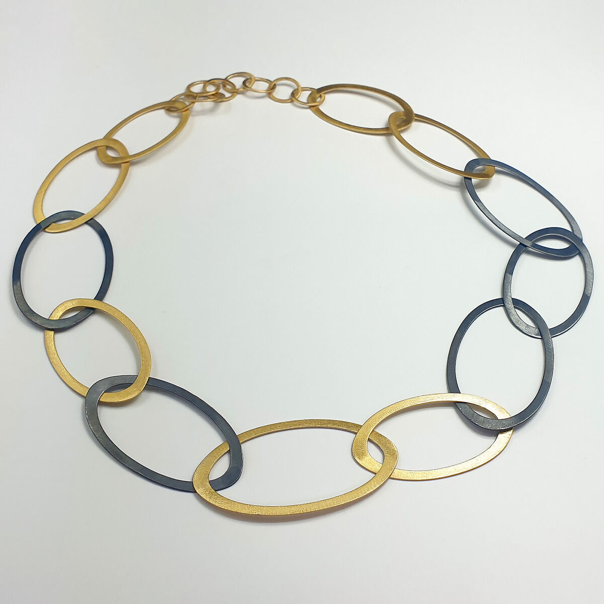 Das Collier »Silberringe« von Maria Rzewuska besteht aus großen, ineinandergreifenden Ringen aus teilweise oxidiertem 925er Silber und feingoldplattierten Elementen. Die Kette wirkt modern und edel durch den Wechsel aus goldenen und dunkelgrauen Ringen auf weißem Grund.