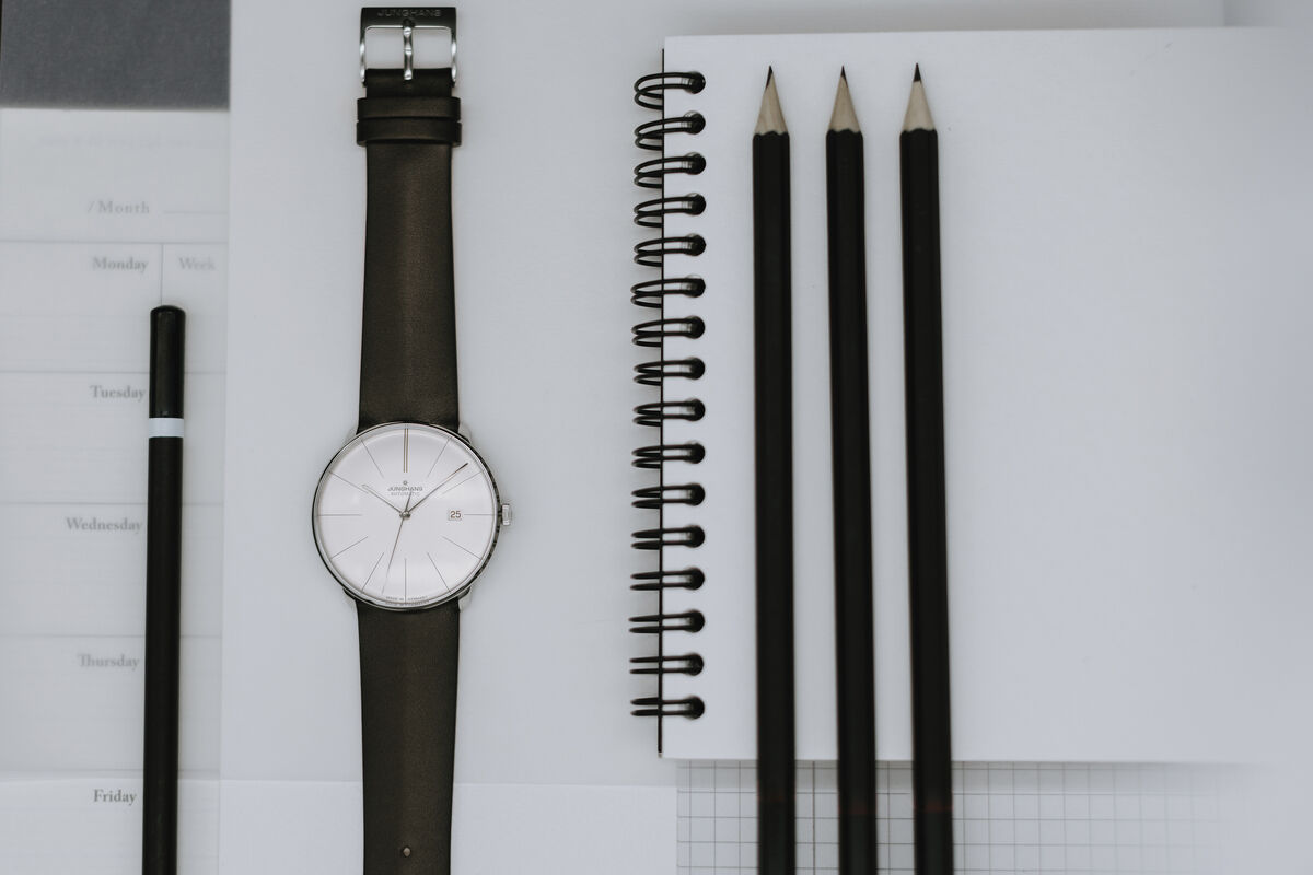 Ein stilvolles Arrangement einer Junghans ZEIT-Sonderedition »Meister fein Automatic« Armbanduhr mit einem schwarzen Lederarmband. Die Uhr befindet sich neben einem Notizbuch, Schreibstiften und einem Kugelschreiber. Die Szene ist minimalistisch und elegant in Schwarz und Weiß gestaltet.