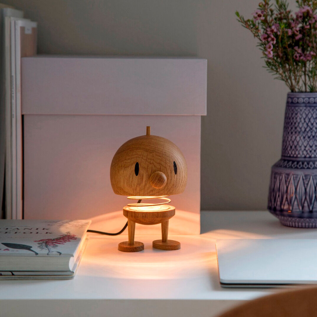 Hoptimist Lampe L aus Eiche auf einem Tisch, mit einem verspielten Design in Form eines kleinen Charakters, umgeben von weißen und pastellfarbenen Objekten.